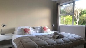 A neatly made bed with a brown quilt, white pillows, and a red decorative pillow. There are two bedside tables with lamps, a window with a view of greenery, and a plain wall. - Private Island Eco Retreat complete with resident Kiwi (Kerikeri)