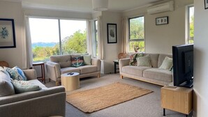 A spacious living room with two sofas, a coffee table, and a television. Large windows provide a view of the outdoors. - Private Island Eco Retreat complete with resident Kiwi (Kerikeri)