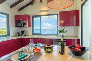 A modern kitchen with red cabinets, a large window with a view, and a dining area set with plates, glasses, and a bottle. - Villa Big Blue, panoramic sea view and pool -5 rooms -Wifi (Le François)