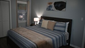 Un dormitorio con cama, mesita de noche, lámpara y una obra de arte en la pared.