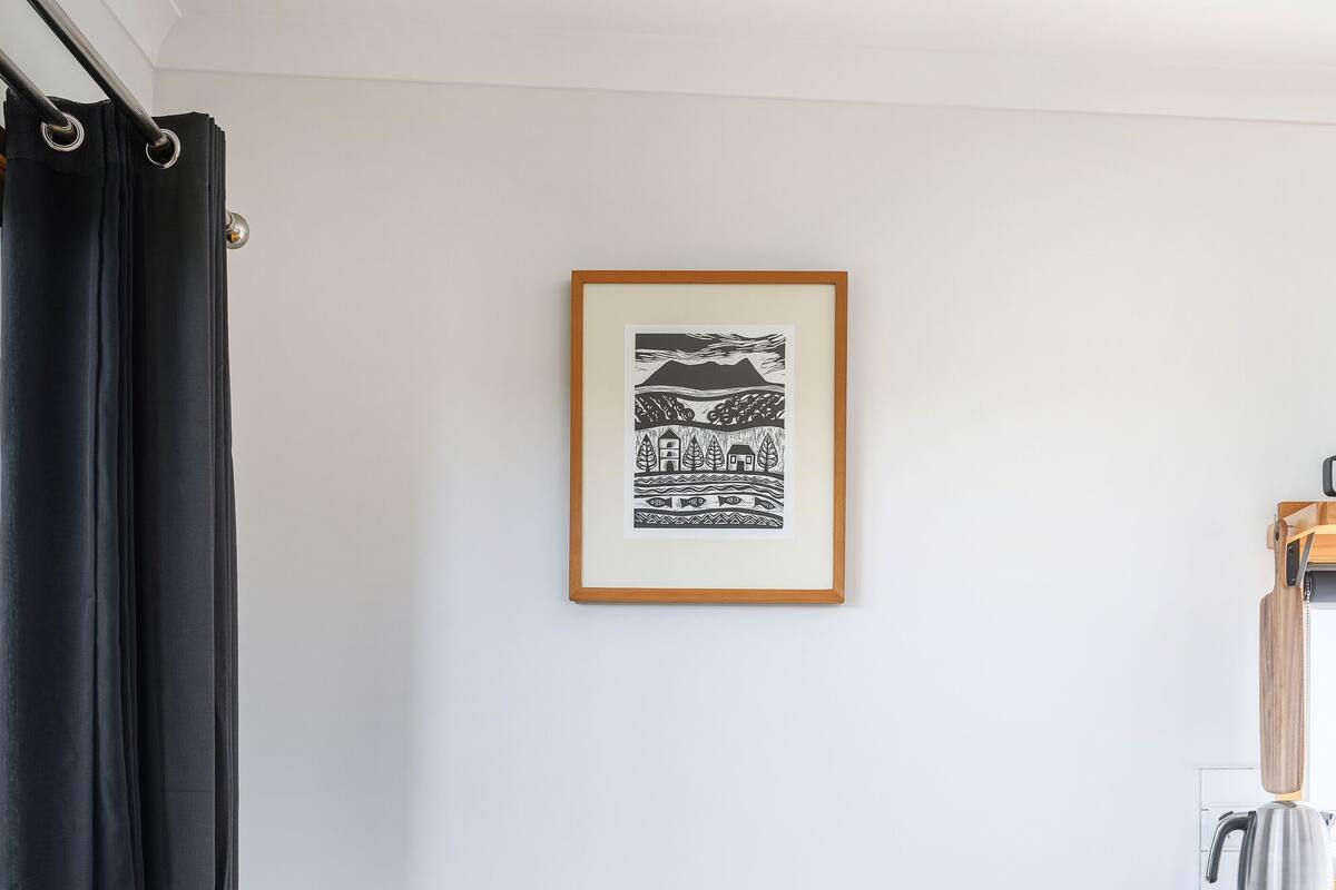 A small dining area with a round wooden table, two black chairs, a framed artwork on the wall, and a flower vase.