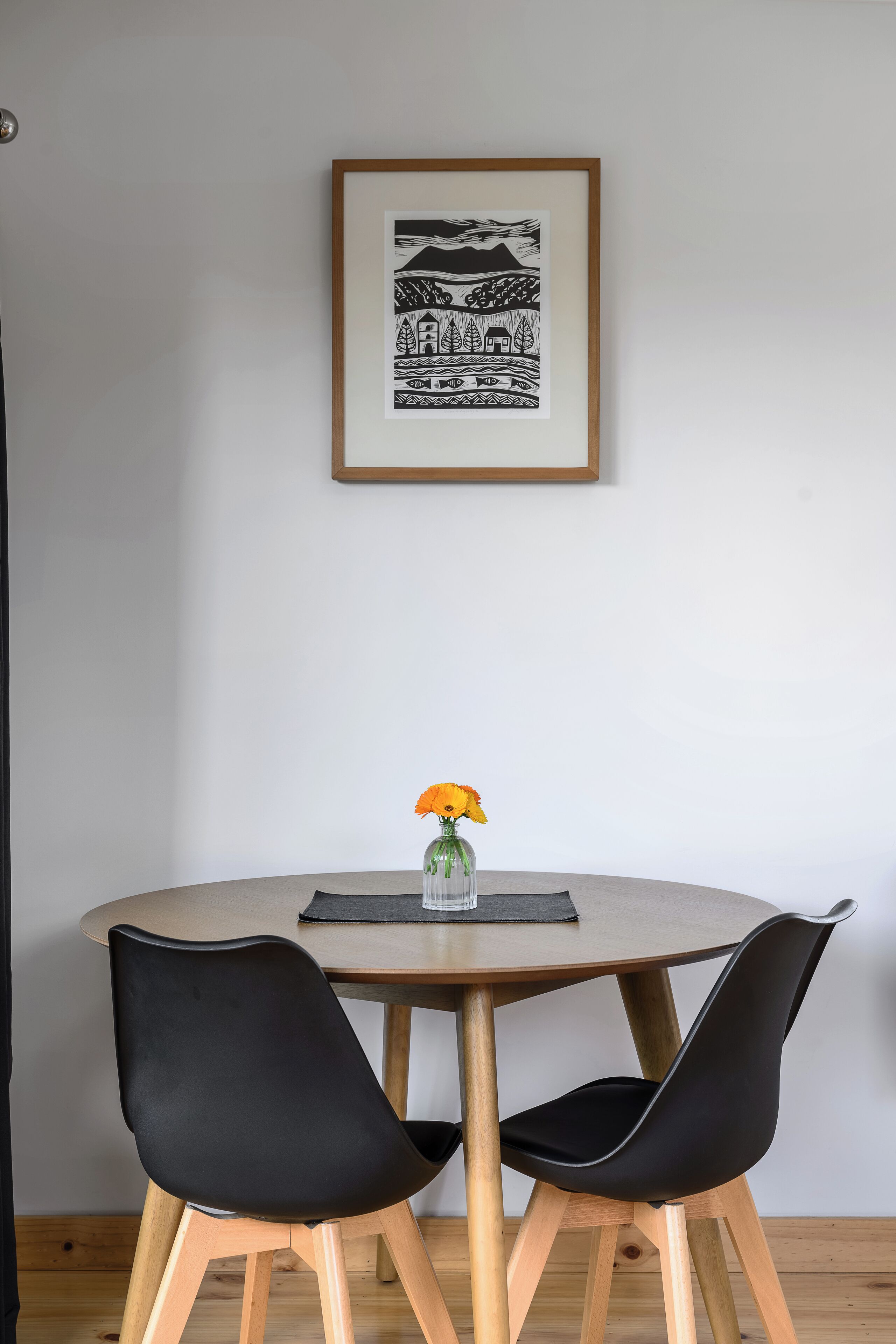 A round dining table with two black chairs, a framed black and white artwork above, and a vase with yellow flowers on the table.