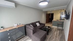 Una sala de estar moderna con un sofá gris, un aparador de madera y una mesa de centro. Se ve una cocina al fondo.
