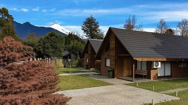 A row of rustic cabins with a wooden deck in front, surrounded by greenery and mountains in the background. - Cabin 1 - Aires del Sur Pucón (Pucon)