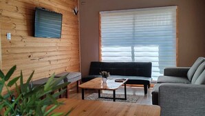 A cozy living room with wooden walls, a flat-screen TV mounted on the wall, a large window with blinds, and a grey sofa. - Cabin 1 - Aires del Sur Pucón (Pucon)