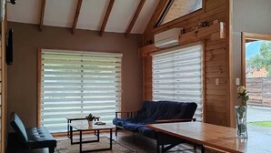 A cozy living room with a sofa, a wooden table, and benches. There are large windows with blinds, a ceiling with wooden beams, and a small potted plant on the table. - Cabin 1 - Aires del Sur Pucón (Pucon)