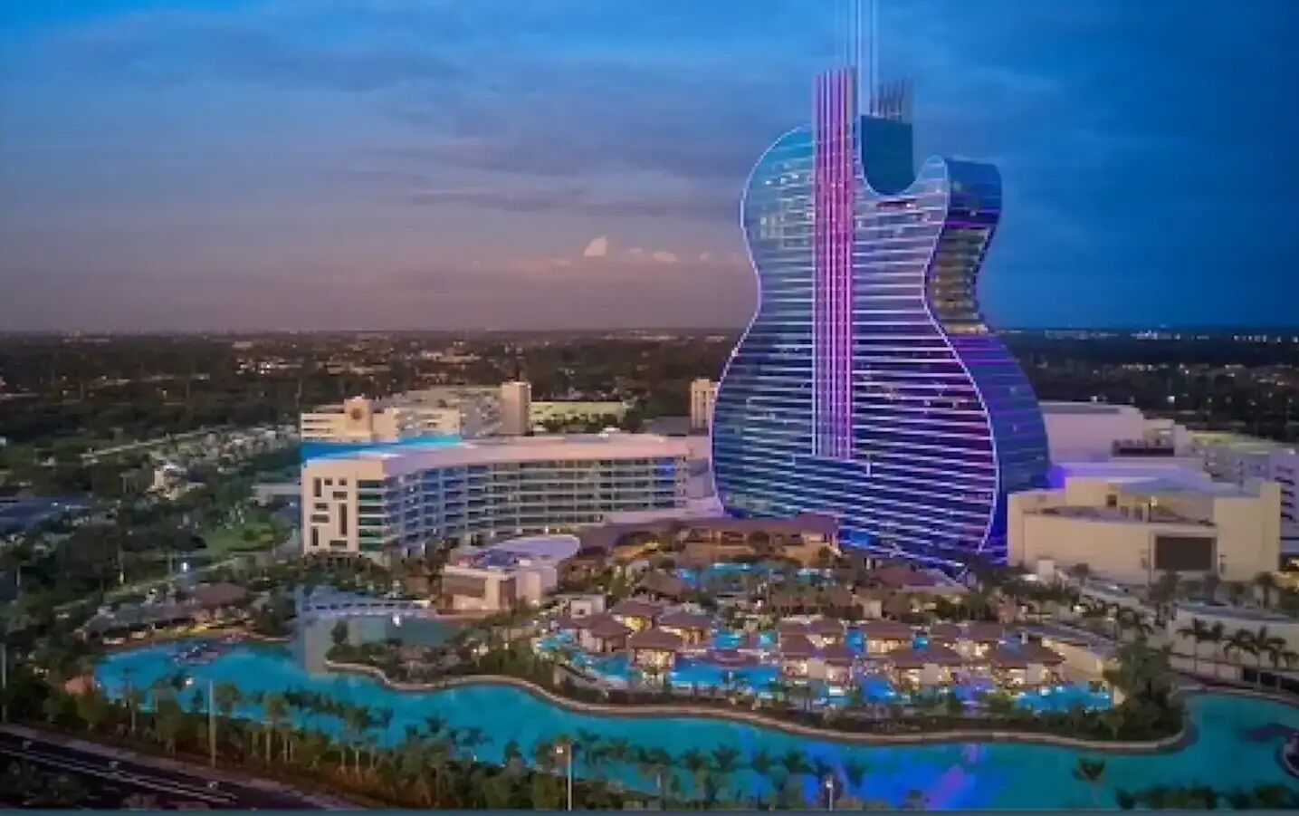 A large resort with a distinctive guitar-shaped building, multiple swimming pools, and surrounding villas.