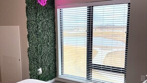 Un dormitorio con cama, ventana con persianas, letrero de neón y un cuadro verde en la pared.