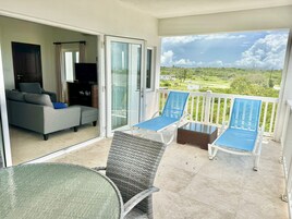 A balcony with a sofa, two blue lounge chairs, a glass table, and a view of a green landscape.