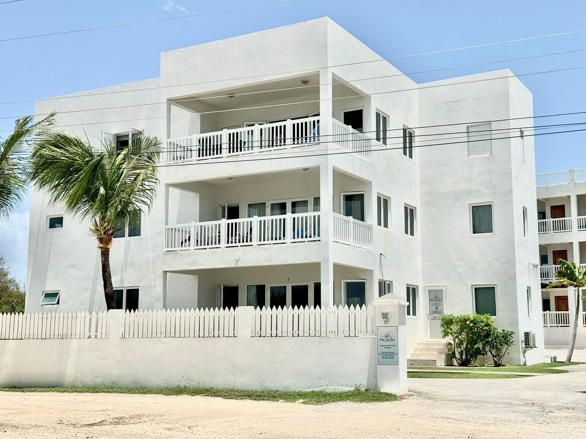 A multi-story building with balconies, a white picket fence, and a palm tree.