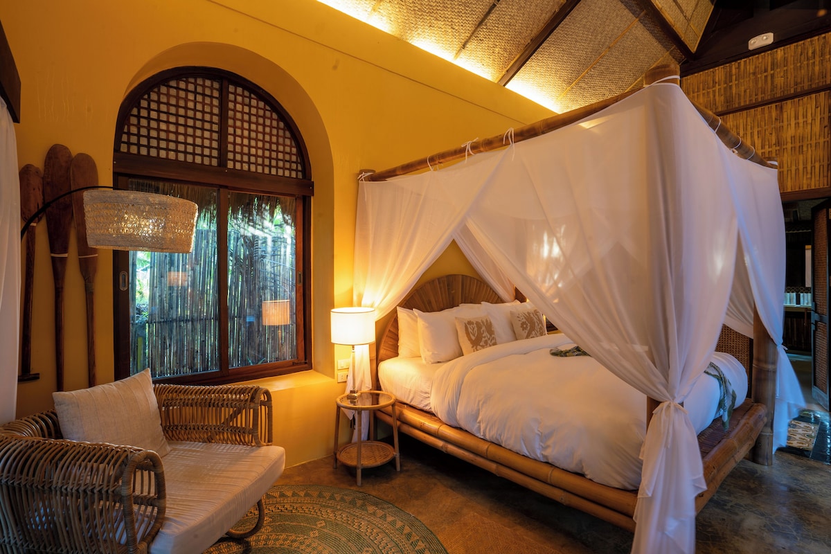 A room with a bed under a canopy, a wicker sofa, a small table with a lamp, and a window with a view of greenery.