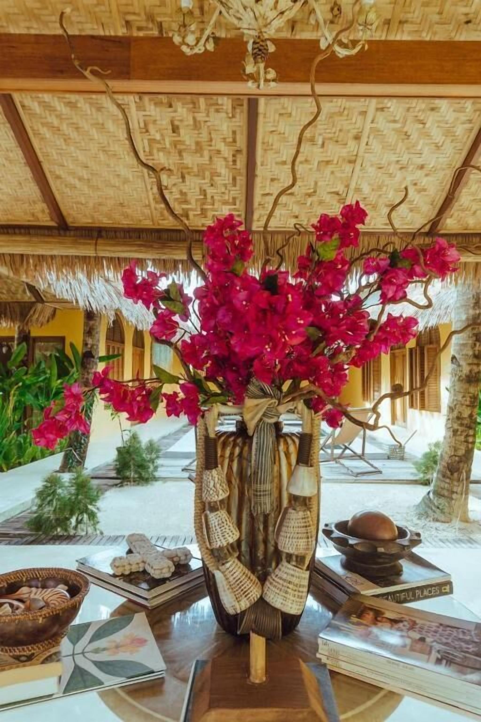 A room with a thatched roof, a wooden table with a vase of red flowers, and a woven basket.