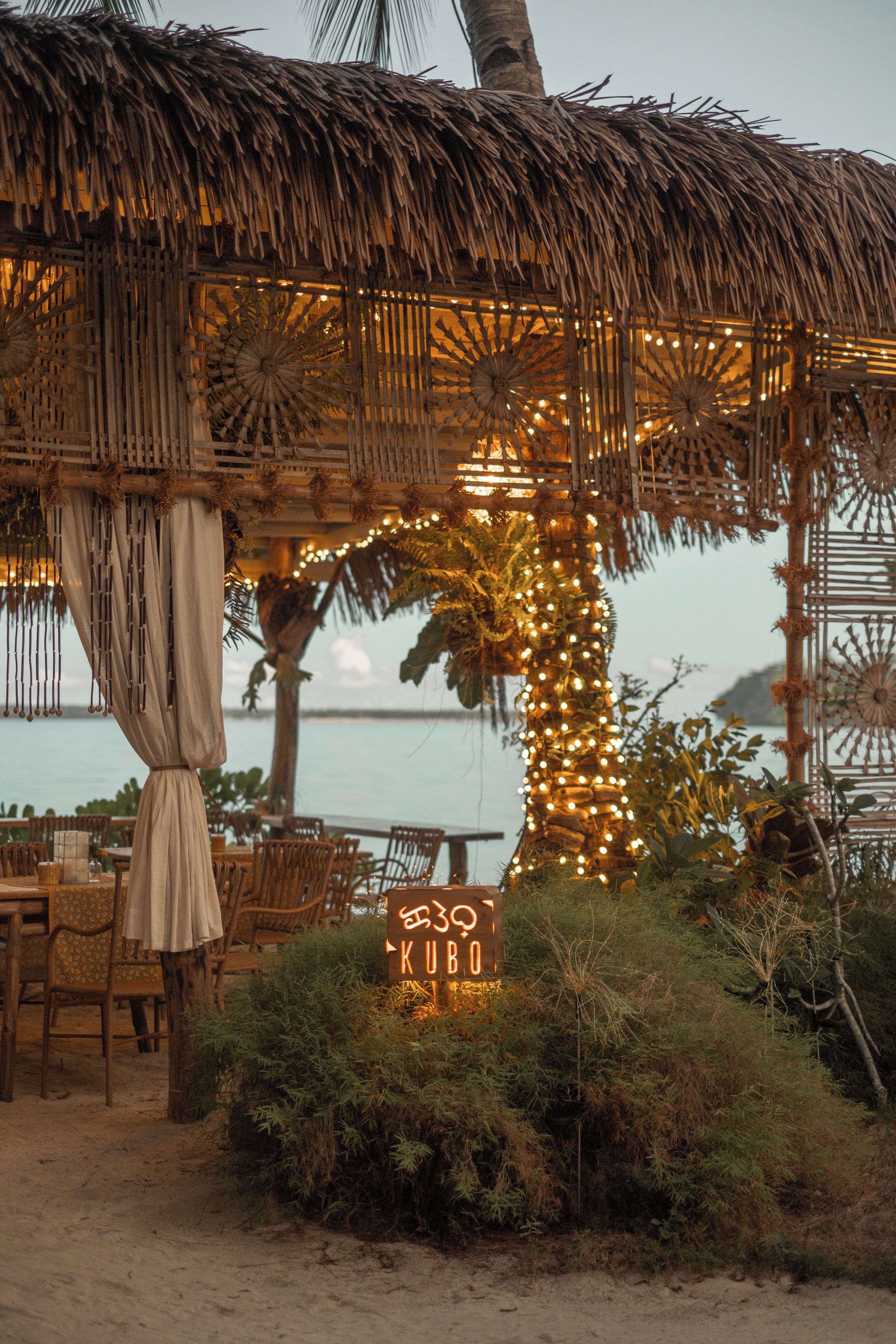 A thatched-roof outdoor seating area with string lights, a sign with illuminated letters, and a view of the sea.