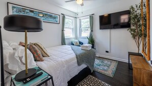 Un dormitorio con cama, ventilador de techo, televisor, ventana con cortinas, una planta y un banco de madera.
