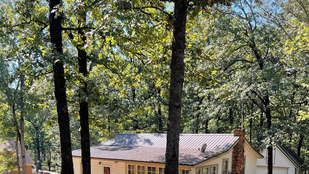 A single-story house with a sloped roof, surrounded by trees and a dirt path.