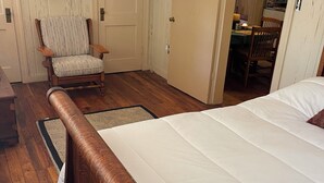 A bedroom with a wooden bed, a chair, a door, and a dining area visible through an open door.