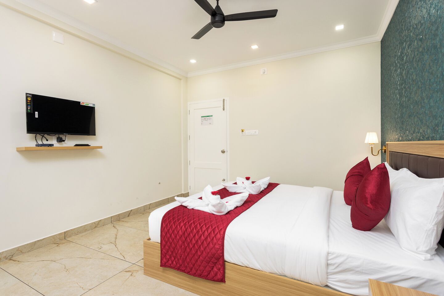 Una habitación de hotel con una cama, un televisor en un estante, un ventilador de techo y una puerta.