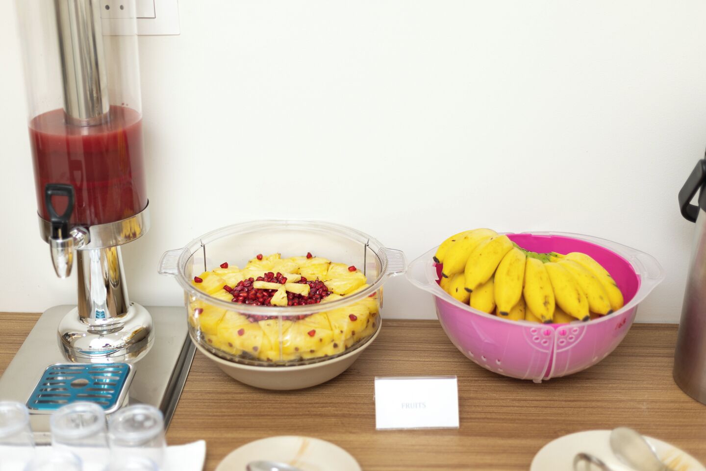 A buffet table with a fruit display including a bowl of bananas, a bowl of pineapple with pomegranate seeds, and a juice dispenser.
