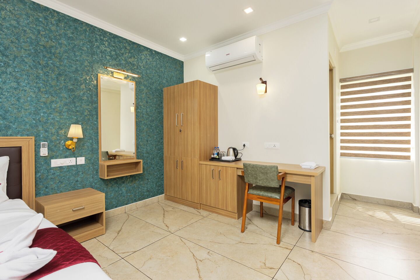 A hotel room with a bed, a desk, a chair, a wardrobe, a mirror, a wall-mounted air conditioner, and a window with blinds.