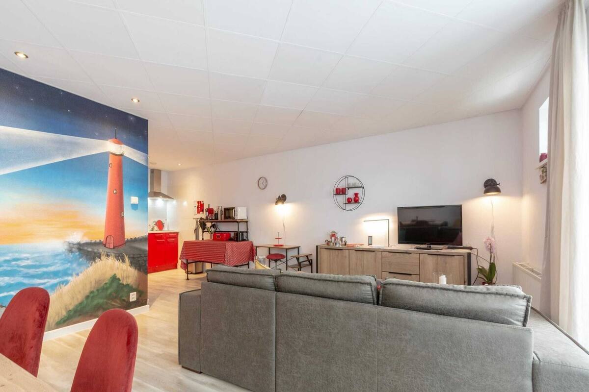 A living room with a large grey sofa, a red dining table, and a kitchen area with a red cabinet and a microwave.