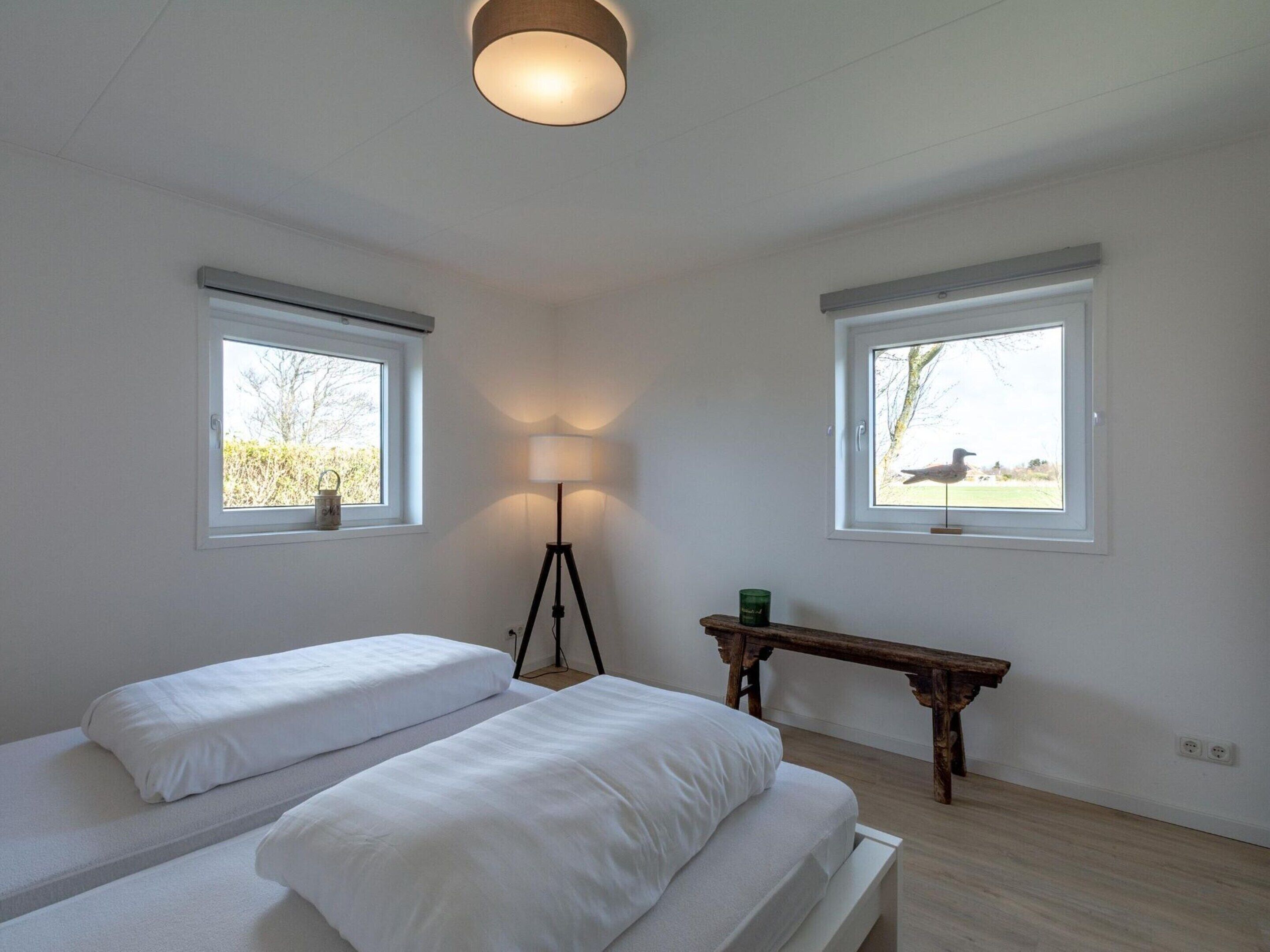 A bedroom with two single beds, a bedside table, a floor lamp, and two windows with views of the outdoors.