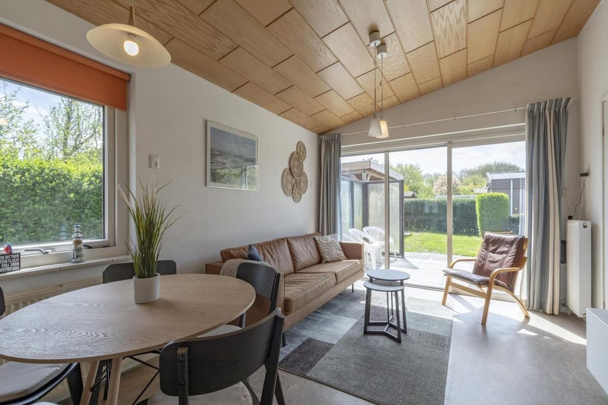 A spacious living area with a wooden ceiling, a round dining table with chairs, a sofa, and a small side table.