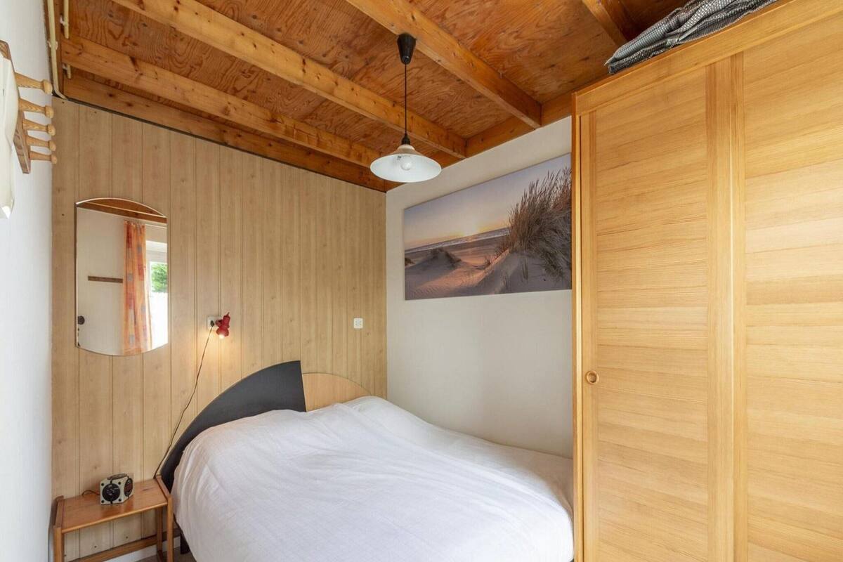 A bedroom with a wooden ceiling, a bed with white bedding, a nightstand with a radio, a mirror, and a wall-mounted picture.