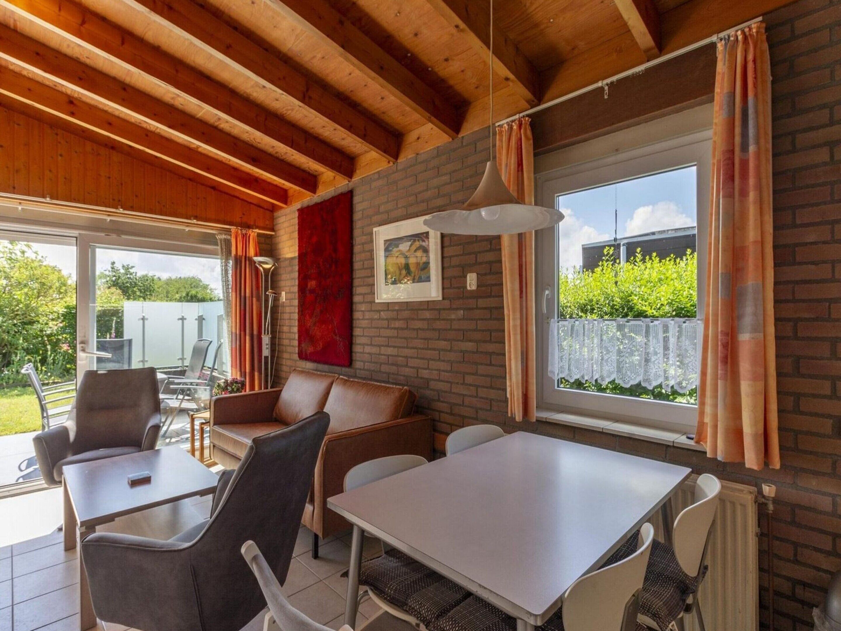 A cozy indoor space with a brick wall, a dining table, and chairs. There is a window with curtains, a hanging light fixture, and a red abstract painting.
