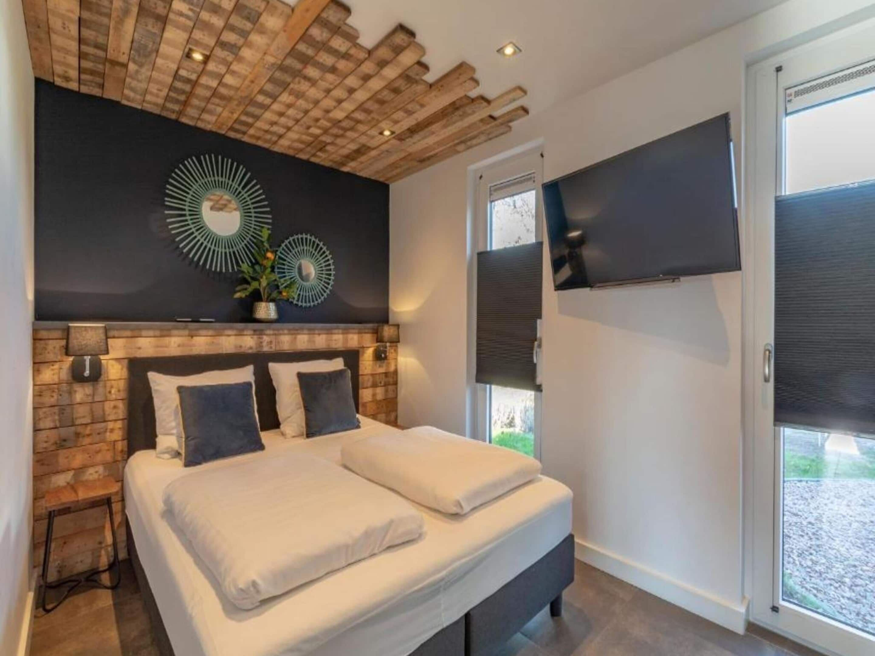 A modern bedroom with a brick-patterned headboard, a large bed with white linens, two decorative wall mirrors, and a large window with a view of the outdoors.