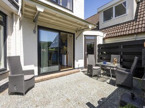 A patio with outdoor furniture, a sliding glass door leading to a kitchen, and a small garden area. - Detached 6 Pers Holiday Home (De Koog)