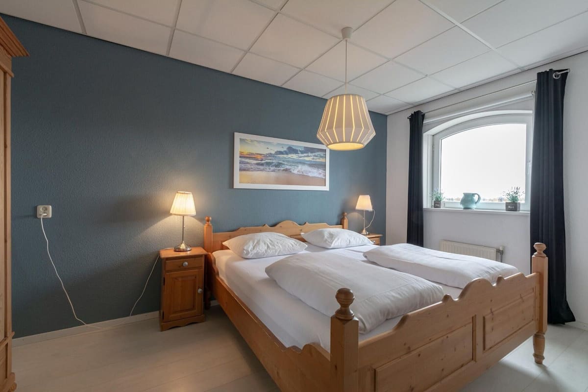 A bedroom with two beds, a wooden headboard, bedside lamps, a painting of a beach, and a window with curtains.