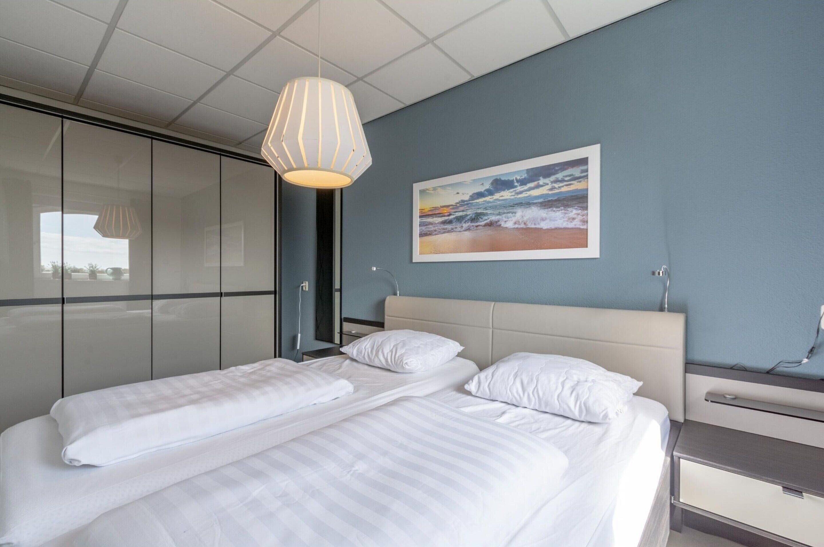 A hotel room with a bed, a bedside table, a lamp, and a painting of a beach scene.