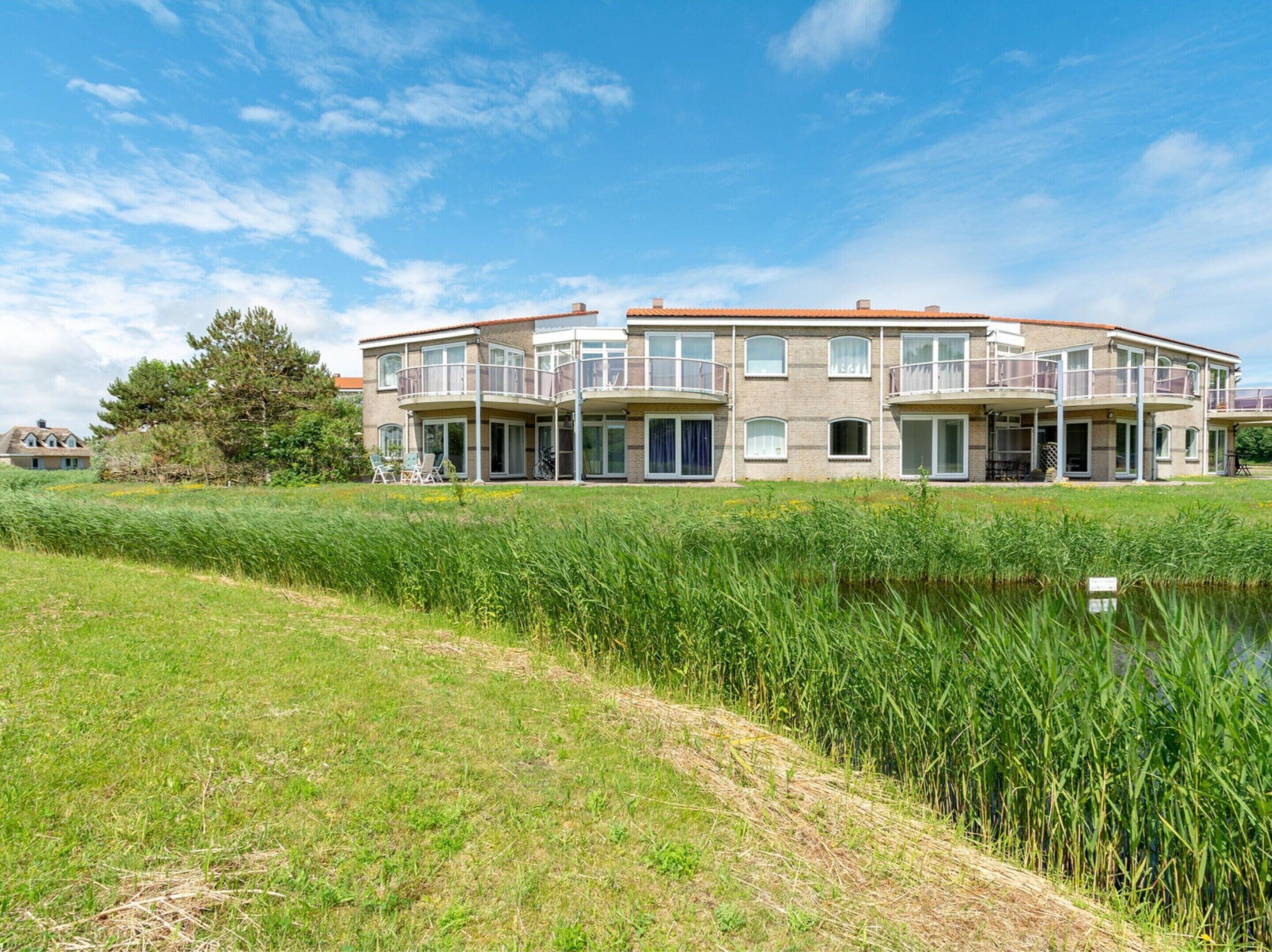 A modern multi-story residential building with balconies, a grassy foreground, and a body of water.