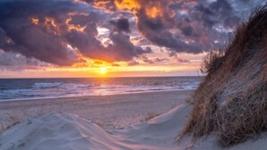 Ein Strand mit einem Weg zum Meer, einer Düne und einem Sonnenuntergang im Hintergrund.