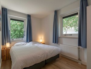A bedroom with a bed, two bedside tables, a window with curtains, a radiator, and a view of greenery outside.