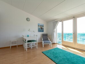 A room with a white table and chairs, a grey armchair, a green rug, a painting of a boat on water, and a large window with a view of the sea. - The Lovely Apartment is Beautifully Decorated (Julianadorp)