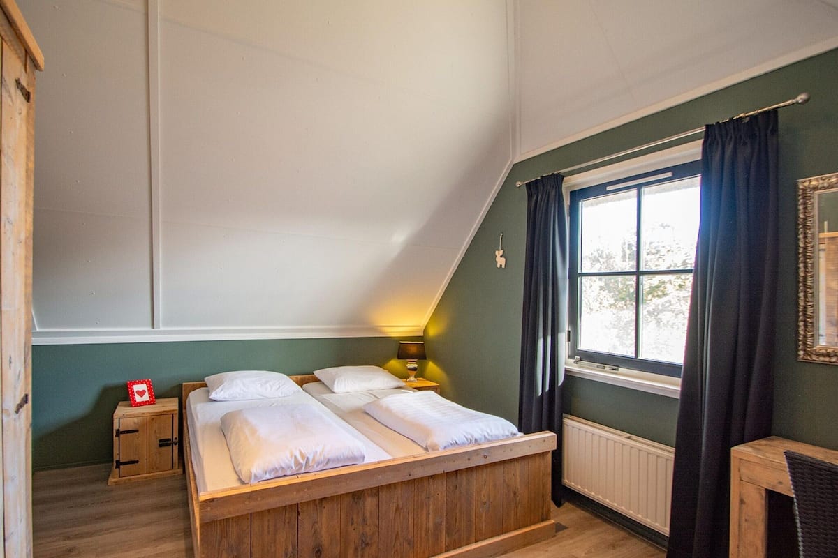 A bedroom with a sloped ceiling, a wooden bed, a bedside table with a lamp, a window with curtains, and a radiator.