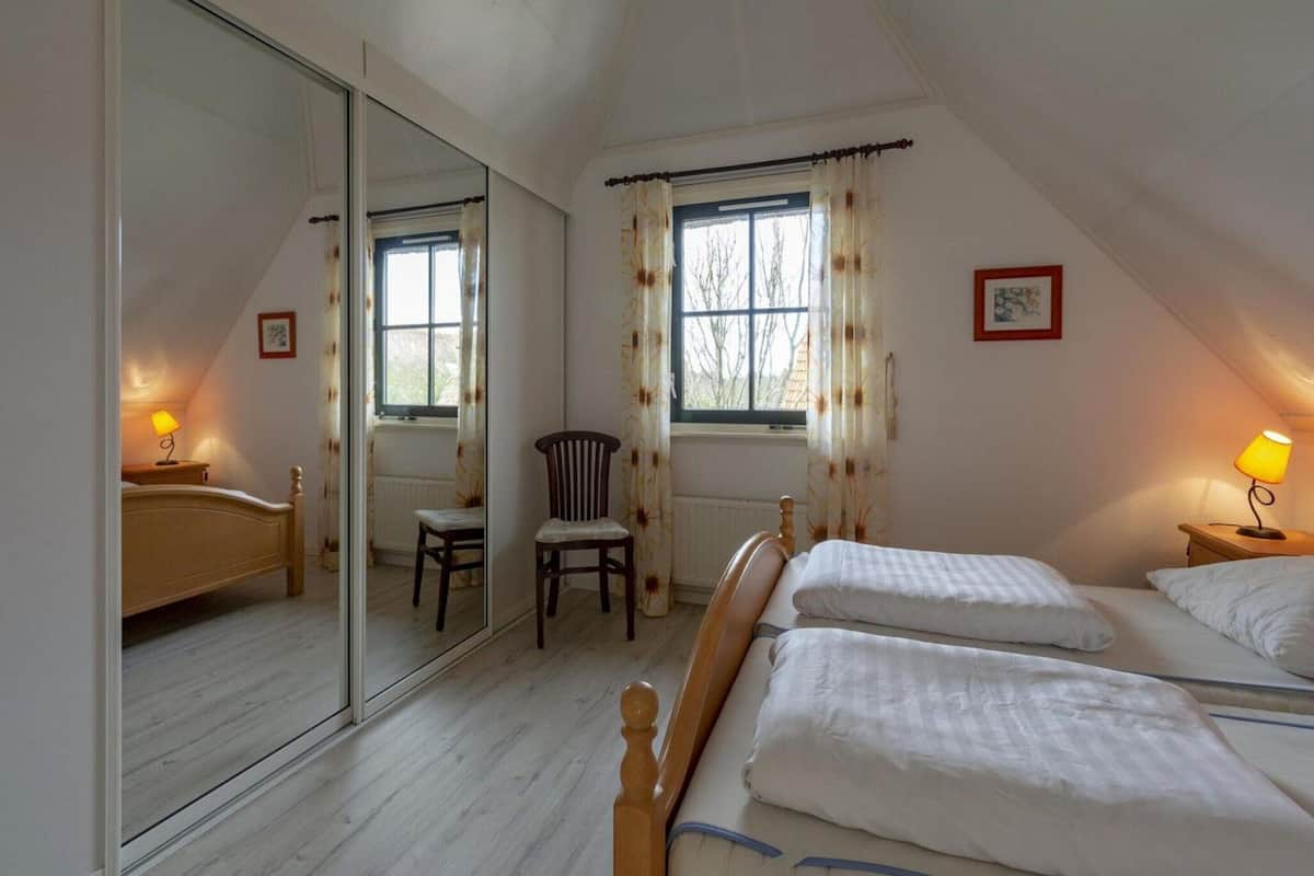 A bedroom with two beds, a chair, a window with curtains, a radiator, and a mirror.