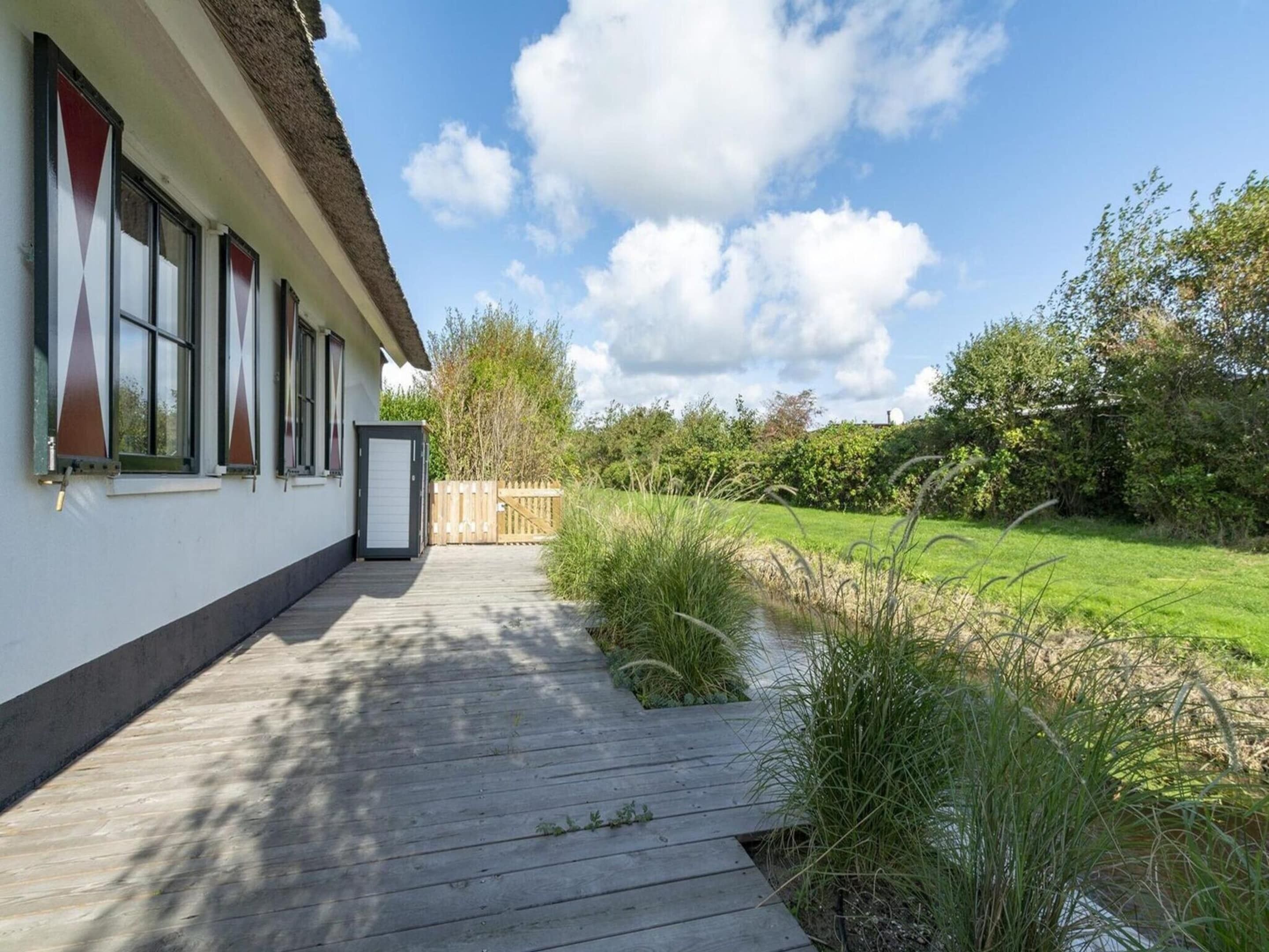 A wooden deck leading to a house with a thatched roof, a small pond, and a grassy area with trees.