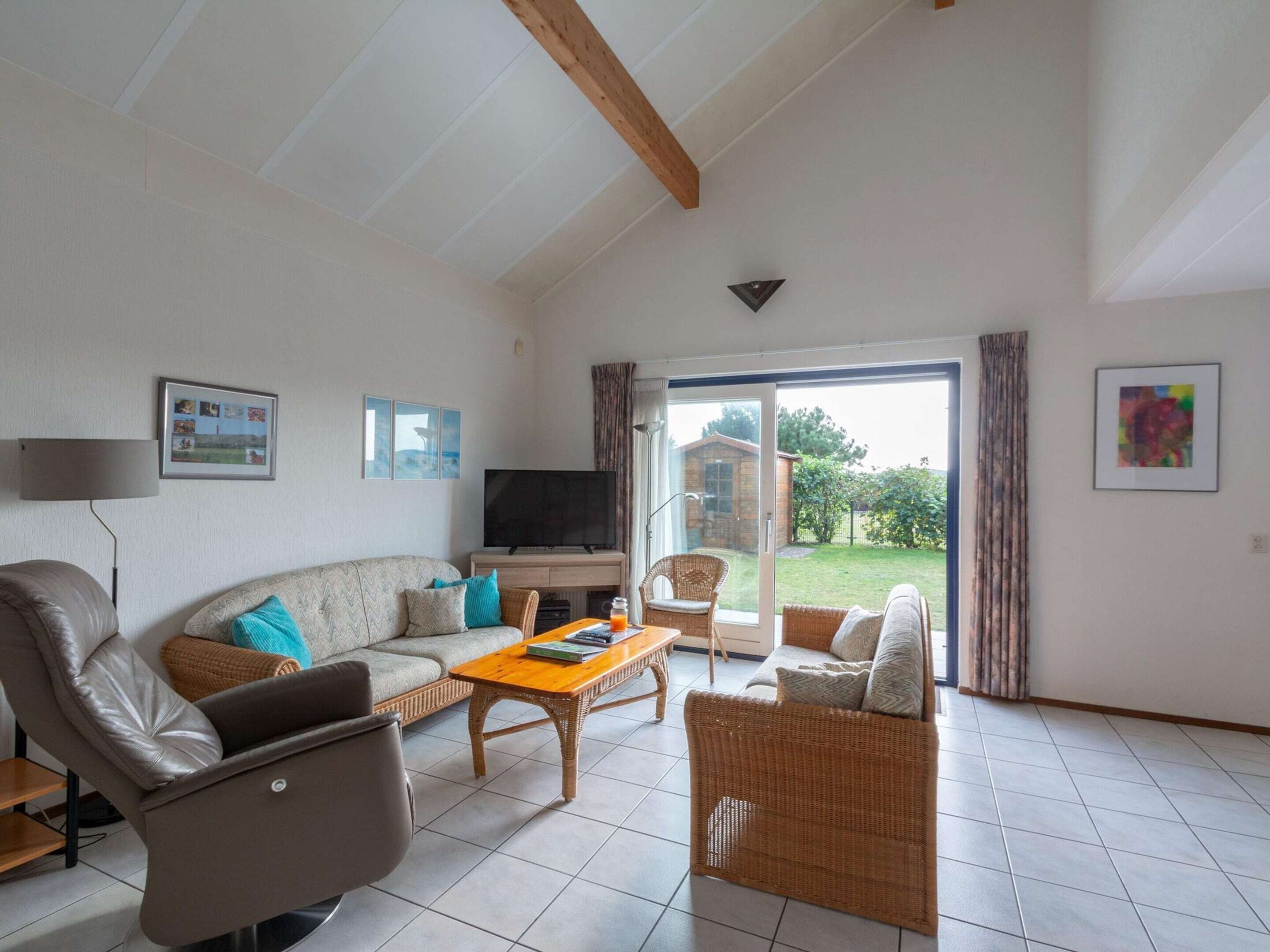 A living room with a sofa, armchair, and wicker chairs, a coffee table, a television, and a sliding glass door leading to a garden.