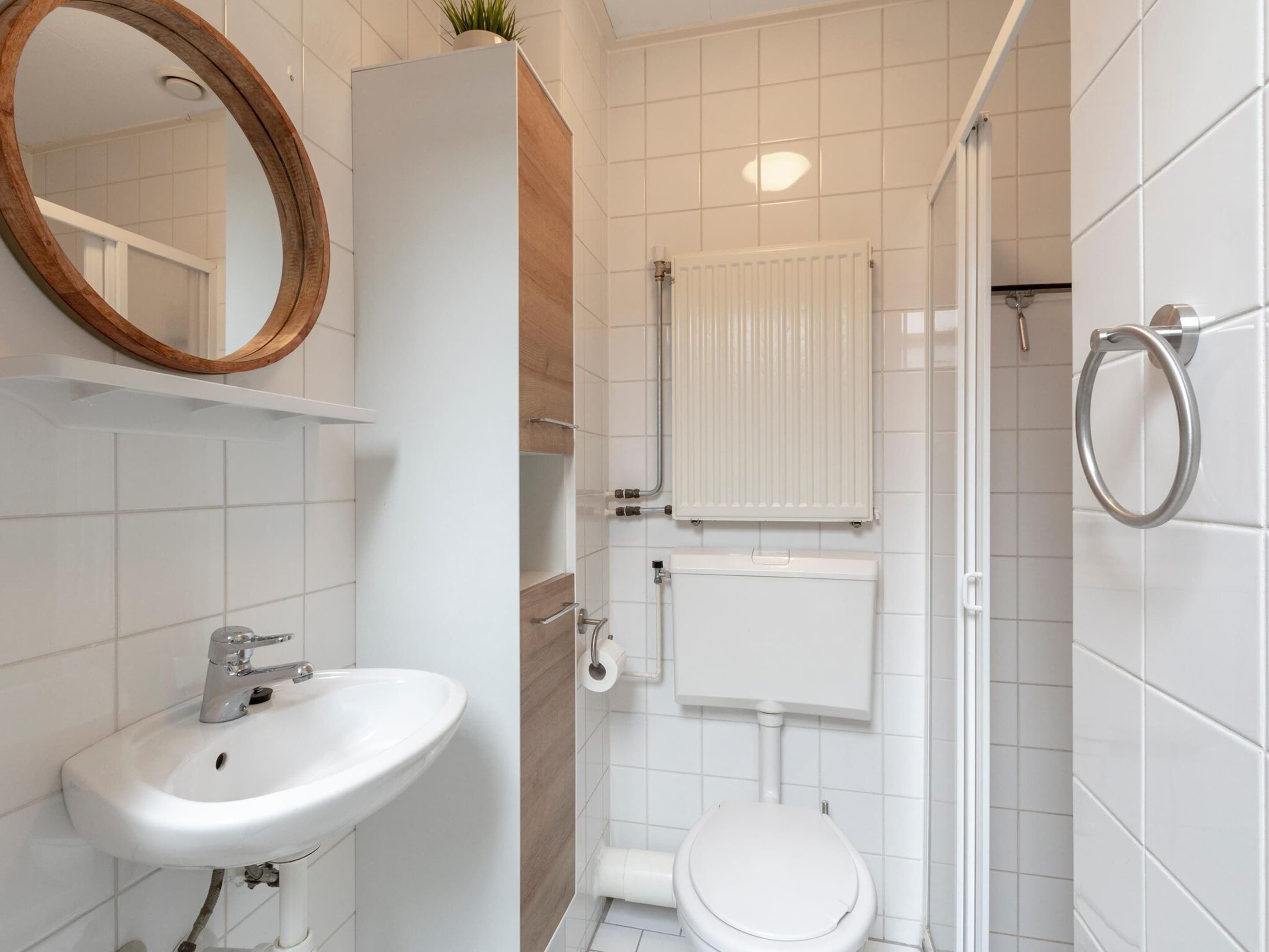 Un baño con un lavamanos, inodoro y ducha blancos. Hay un espejo sobre el lavamanos y un radiador sobre el inodoro.
