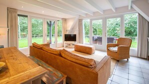 Amplia sala de estar con mesa de comedor de madera, un sofá y un sillón de mimbre. Hay un televisor sobre un soporte blanco y un gran ventanal con vistas al exterior.