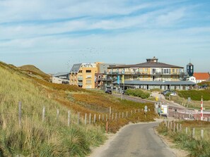 A coastal road leading to a multi-story building with balconies, a lighthouse, and a beach in the background.