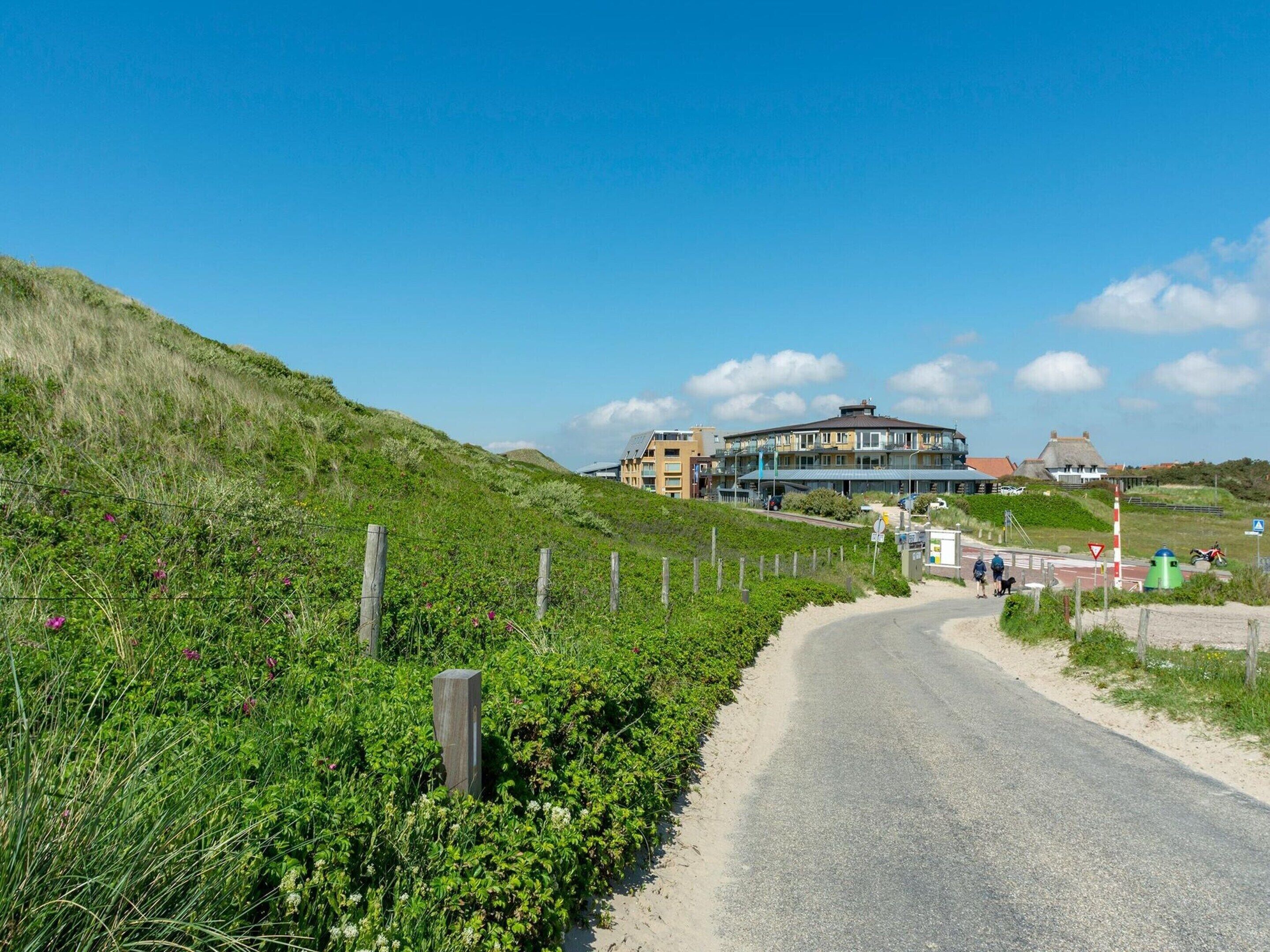 A coastal road with a building complex in the background, surrounded by greenery and a clear sky.