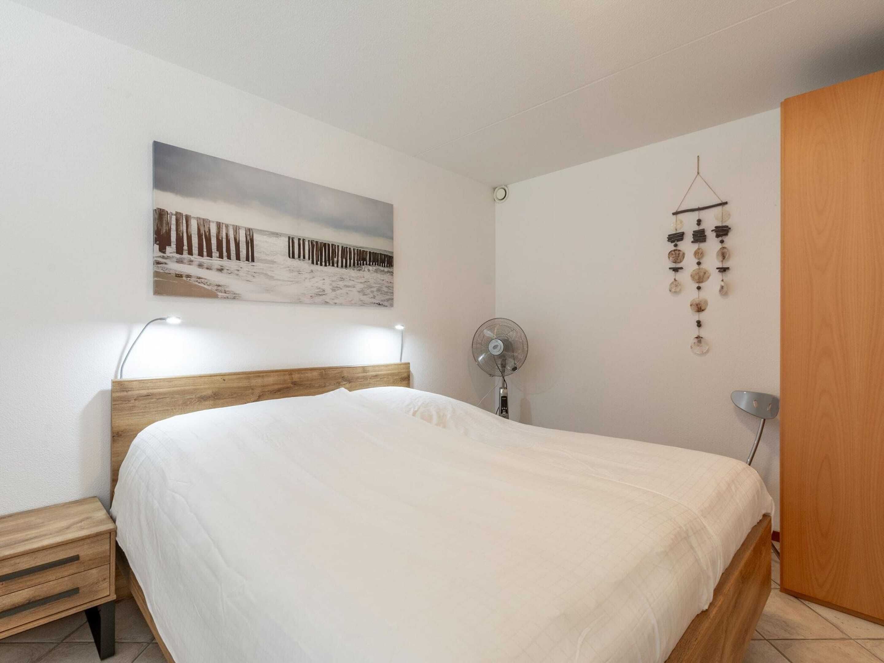 A bedroom with a wooden bed, a bedside table, a wall-mounted fan, and a painting of a snowy landscape.