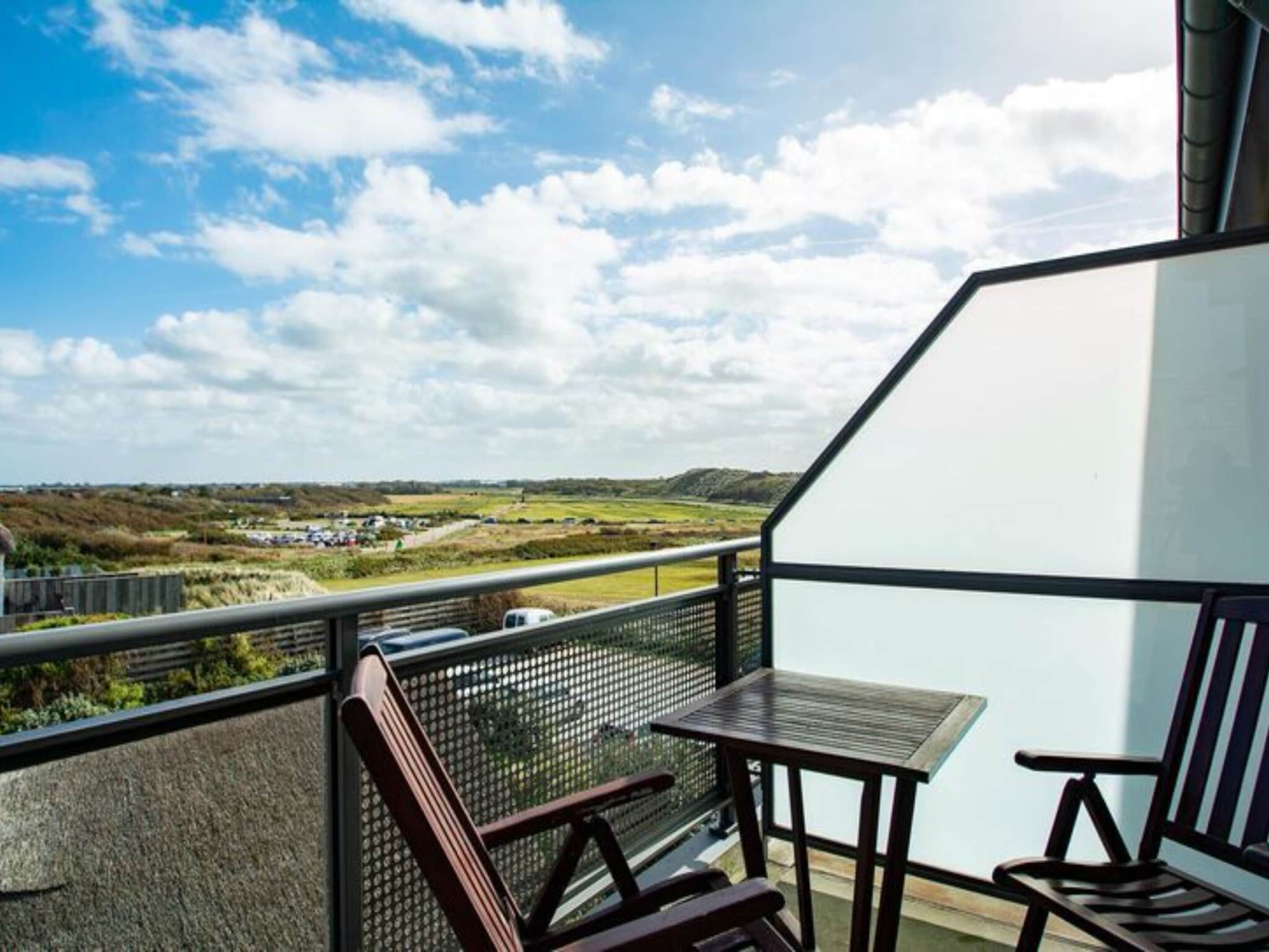 A balcony with a table and chairs, overlooking a landscape with fields and distant buildings.