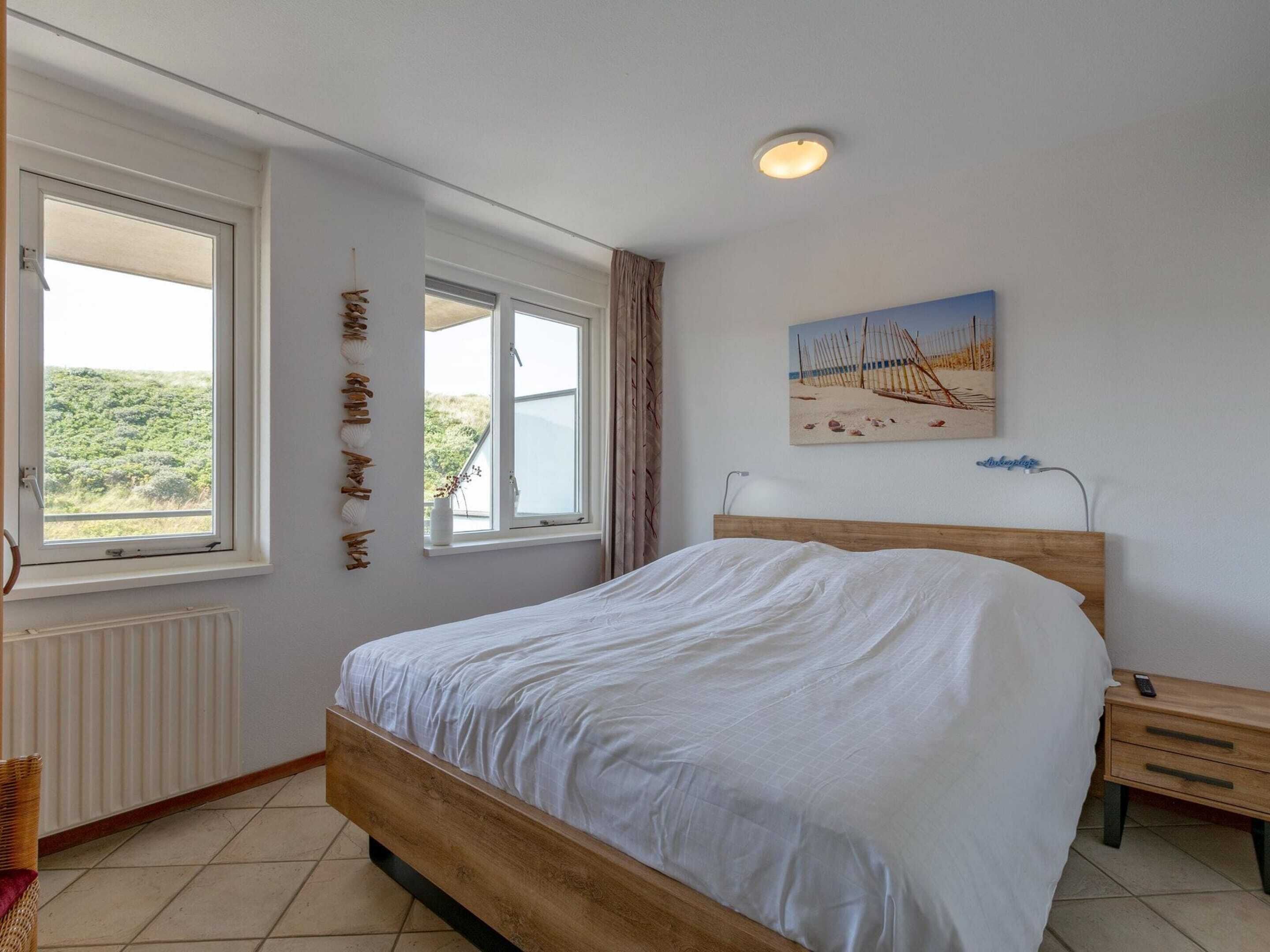 Ein Schlafzimmer mit einem hölzernen Bett, zwei Fenstern, einer Heizung, einem Nachttisch und einem Strandmotiv-Bild.