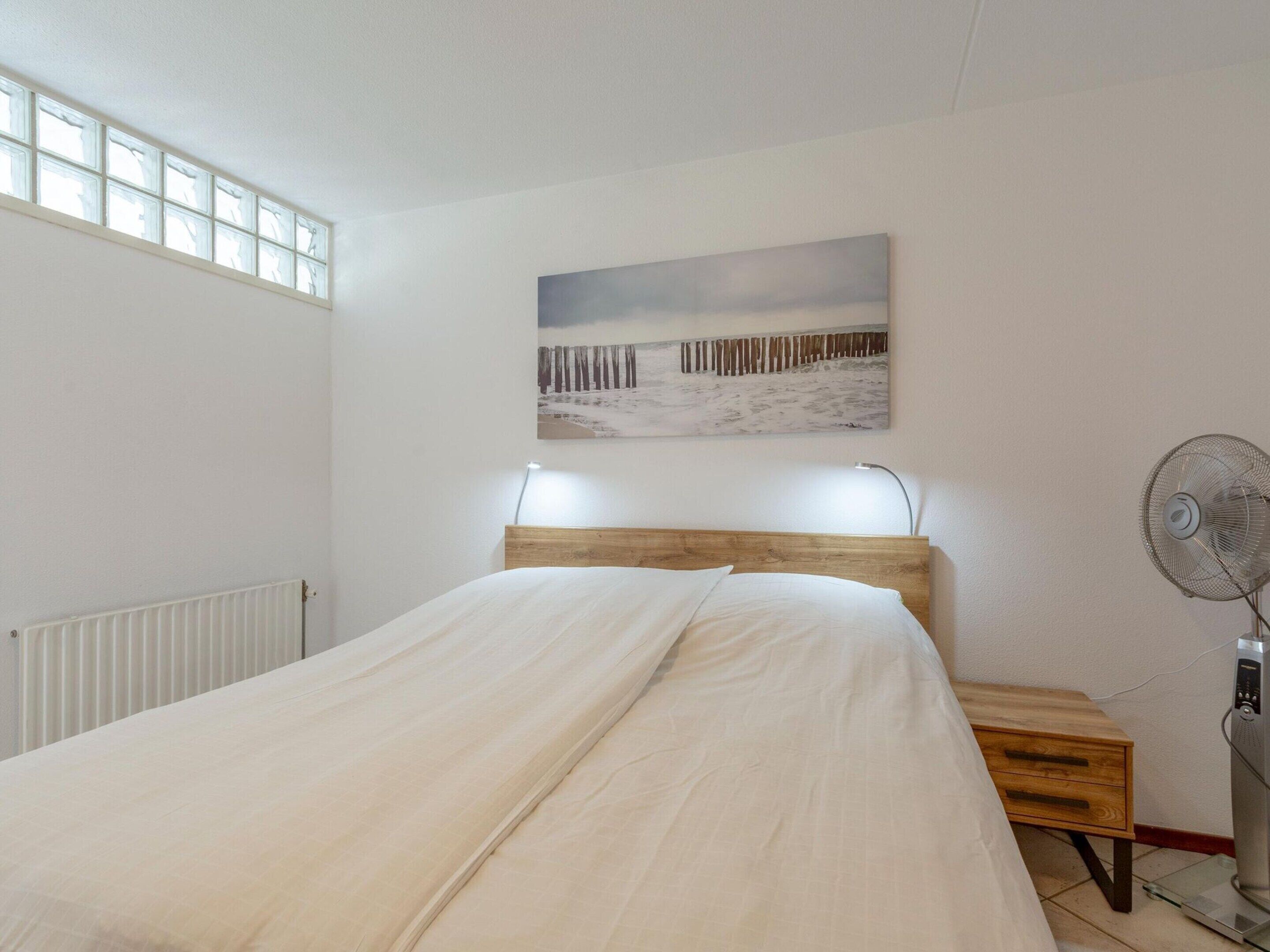 A bedroom with a bed, a bedside table, a standing fan, a radiator, and a window.