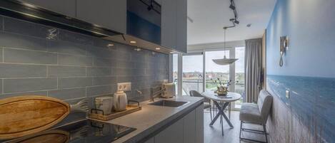 Eine moderne Küche mit einer großen Kochinsel, ein Essbereich mit Blick und ein Wohnbereich mit großem Fenster zum Meer.