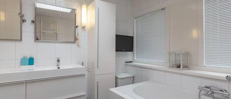A modern bathroom with a large bathtub, a double sink vanity, a mirror, and a window with blinds.